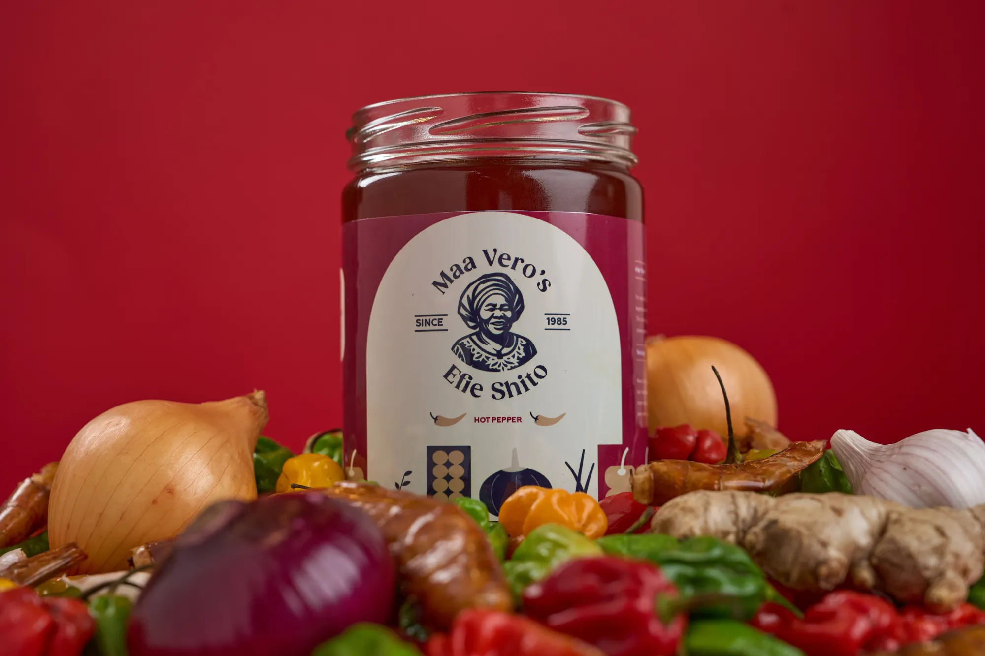A jar of Maa Vero's Efie Shito surrounded by fresh peppers, onions, ginger and garlic